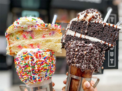 Two Bowery milkshakes, one with rainbow sprinkles and cake on top, and another with mini chocolate chips and chocolate cake on top.