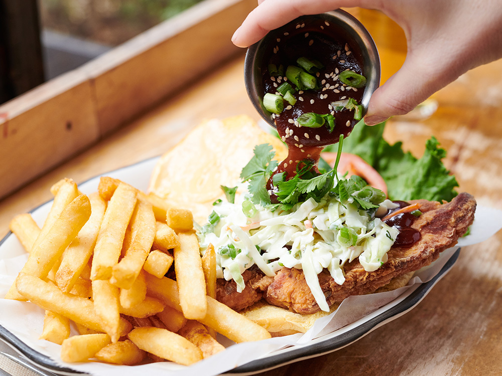 Sauce being poured on a Chicken Sandwich with a side of French fries, one of our many Bowery, New York City food delivery options.